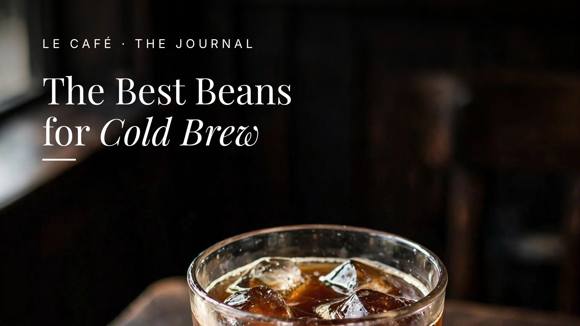 A glass of cold brew coffee with ice on a dark wood table with whole coffee beans. Le Café Journal header: The Best Beans for Cold Brew