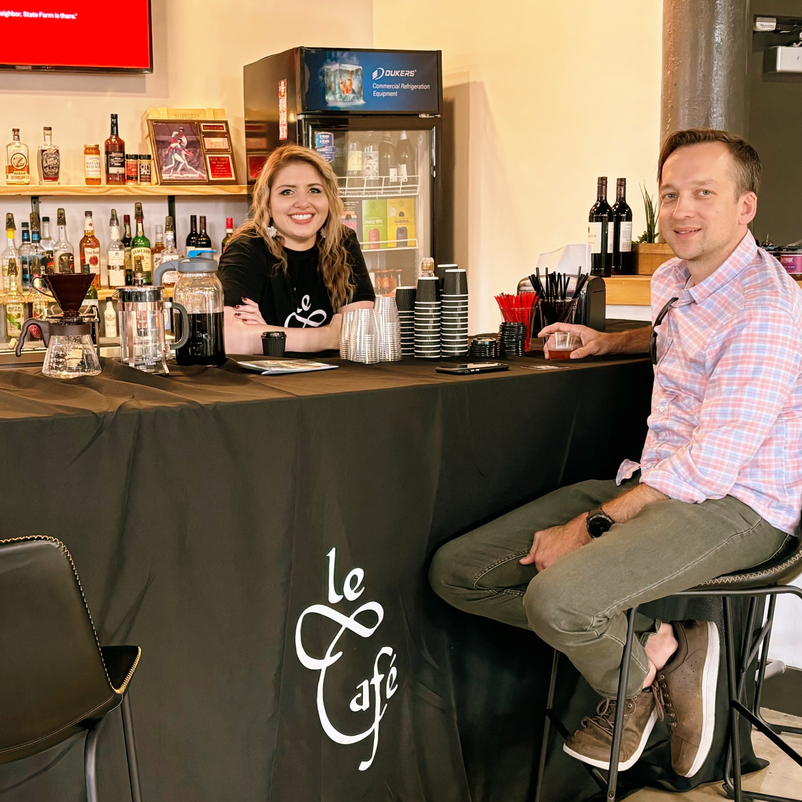 Nechama Henderson smiles while wearing a Le Café shirt stands behind a black table with coffee supplies and branding, serving a male customer seated at a bar-style setup inside a lounge with a visible liquor shelf and refrigerator in the background.