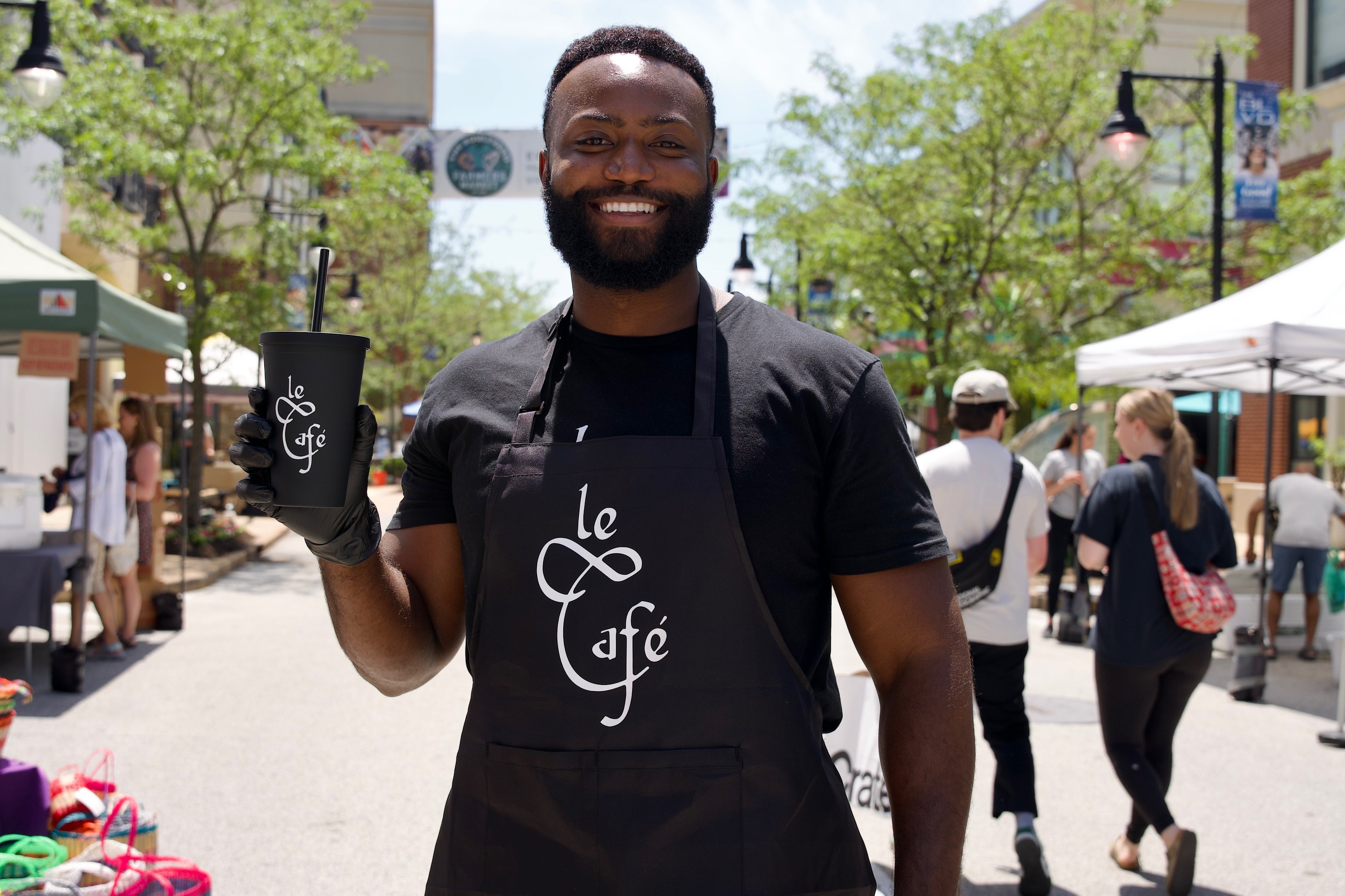 Co Founder Andre Walker wearing a black apron with 'Le Cafe' on it, holding a Le Cafe cup outside of Farmers Market