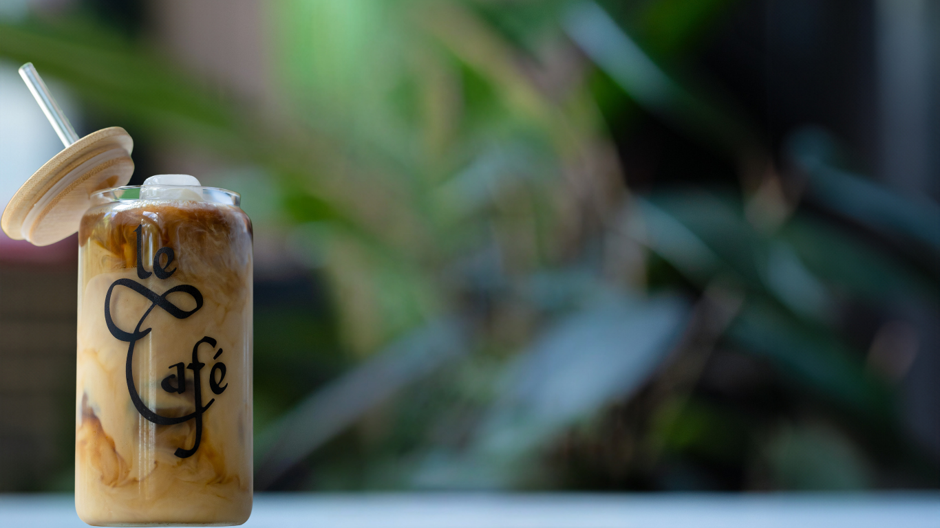 A glass of iced coffee with swirling cream, branded with the elegant black script logo 'Le Café', topped with a bamboo lid and straw, set against a blurred green plant background.