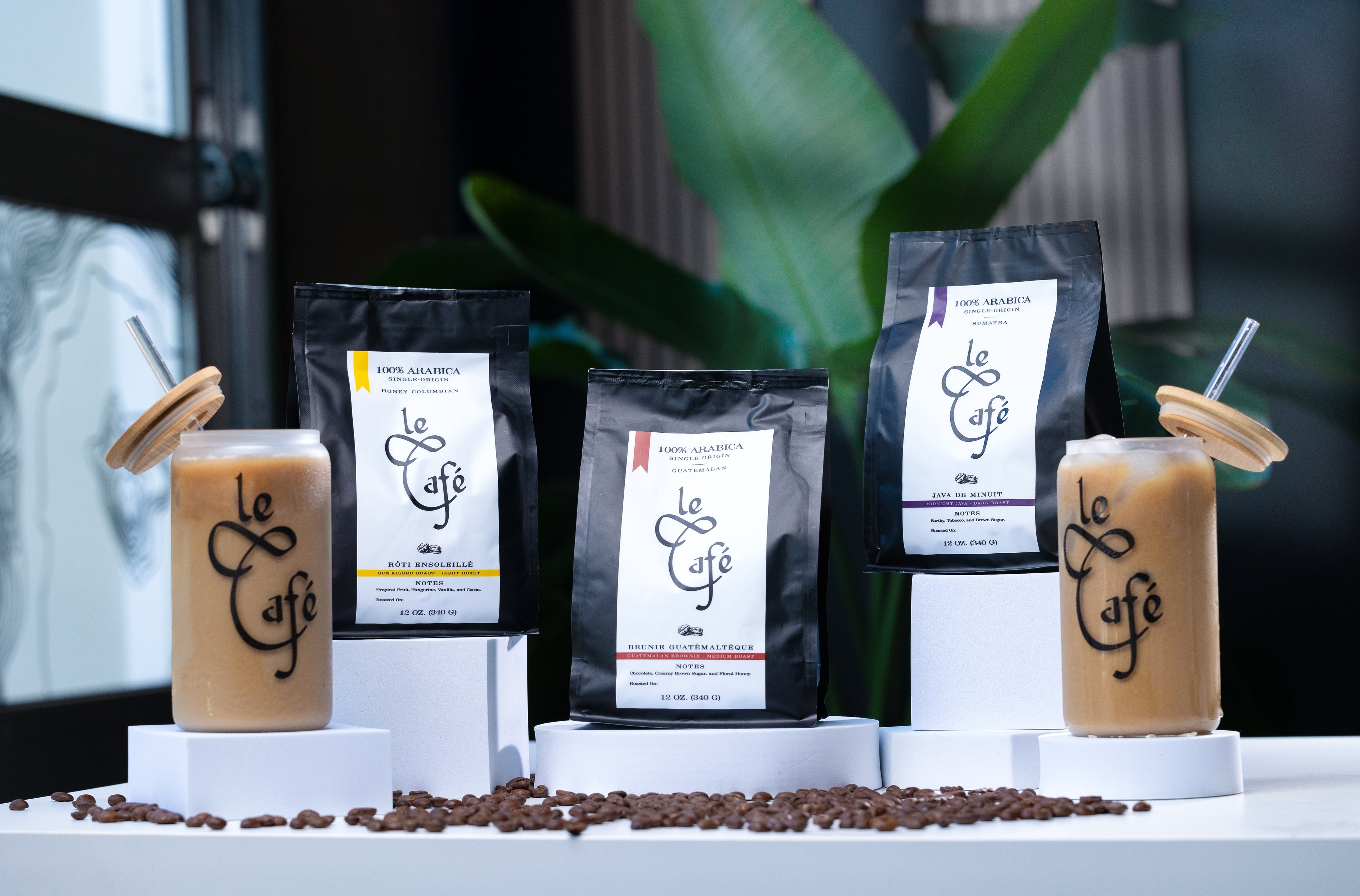 Le Café branded iced coffee drinks in glass jars with bamboo lids and straws, flanking three bags of Le Café 100% Arabica coffee blends labeled from Colombia, Guatemala, and Sumatra, all displayed on white platforms with coffee beans scattered across the surface.